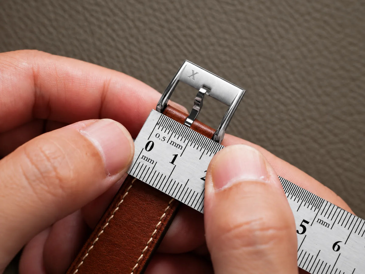 Two hands holding a tan leather strap with its steel buckle, a millimetre ruler laid across the strap just below the buckle frame.