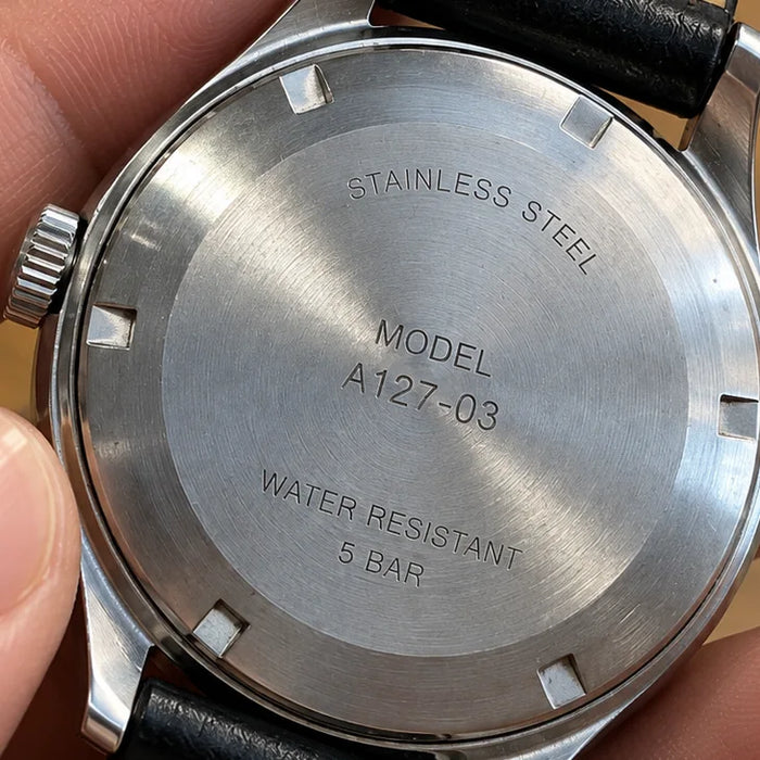 The steel case back of a watch held between two fingers, engraved with its model number.